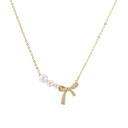 Delicate gold necklace with three white pearls and crystal-embellished bow pendant on cream background