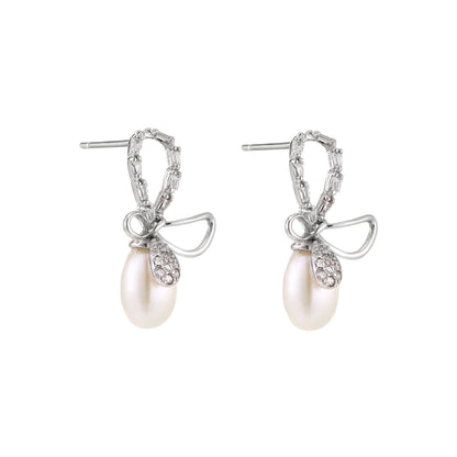 Silver bow-shaped stud earrings with baguette crystals and dangling white freshwater pearls on cream background