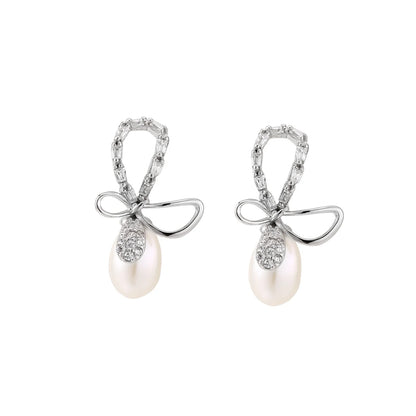 Silver bow-shaped stud earrings with baguette crystals and dangling white freshwater pearls on cream background